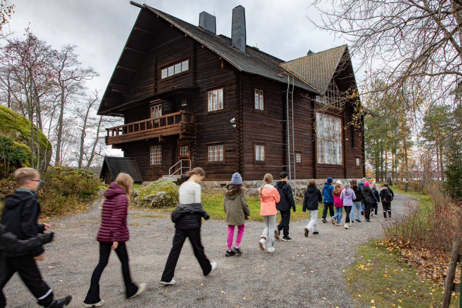 Suuren hirsirakennuksen edustalla kulkee jonossa koululaisia.